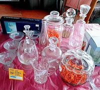 Overview of lot showing various clear glass decanters, stemmed sherbert dishes, a large glass canister with orange flakes and a vintage Pepsi bottle.
