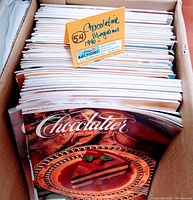 Top view of stacked Chocolatier magazines in box, showing 54 issues from 1990 with cover featuring chocolate layer cake dessert