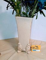 Full view of the cream vase with faux blue and white flower arrangement inside. Vase has a square opening and ornamental leaf feet.