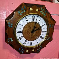 Full frontal view of the round wooden wall clock with scalloped edges, showing the hand painted floral motifs and Roman numeral clock face.