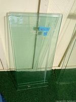 Photo showing multiple green tint glass shelves stacked upright against a wall outdoors, measuring about 26 inches long and 12.5 inches wide, with a label on one piece.