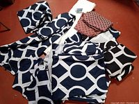 Photo showing multiple folded blue, black, and off-white geometric pattern round tablecloths with two smaller burgundy/gold patterned cloths on top.