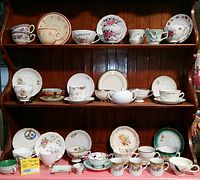 Wide view of a wooden hutch shelf displaying multiple vintage teacups, saucers, plates, and a sugar bowl arranged in rows including items on cup holder stands showing diverse patterns and colors.