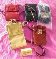 Five Bugatti leather crossbody clutch handbags in red, black, cream, and brown colors laid out on pink surface; includes one sealed purse.