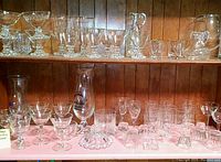 Shelf with multiple rows of vintage stemware glassware including various sizes and designs such as cordial, water, and wine glasses with some crystal and gold rim details.