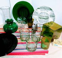 Vintage green glass plates, vases, salt and pepper shakers, cups, glasses arranged on a striped cloth with decorative plate at back.