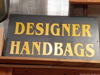 Front view of the black wooden sign with yellow 'DESIGNER HANDBAGS' text.
