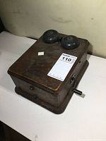 Solid oak antique phone ringer box showing top with two black metal bells and a hand crank on the side, placed on a white surface with aged wood finish.