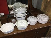 Full set of Corning Ware dishes arranged showing floral pattern stack and white ribbed dishes with lids on wooden surface