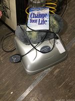 Silver Exerciser 2000 Elite machine base with a gray removable sheepskin cover, digital remote control resting on the machine, and user manual on top.