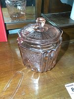 Pink glass biscuit jar with lid standing on wooden surface in front of mirror, showing embossed floral pattern and vertical paneled sides.