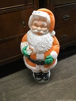 Front view of vintage Santa blow mold with orange coat and white beard, standing on a wooden floor.