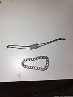 Two silver bracelets on a white surface; one thin charm bracelet with engraved plate monogrammed W.R.C.N.S., one thicker link chain bracelet.