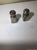 Two sterling silver salt and pepper shakers standing upright on white surface, showing perforated tops and square bodies