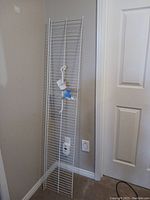 Photo showing the 12" wide wire shelf leaning against a corner wall with a white wall outlet nearby. Shelf is tall and narrow with metal wire construction.