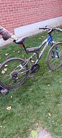 Side view of the entire blue and gray adult mountain bike on grass showing drivetrain, frame, and tires. The front tire and rear tire both appear flat. Hand holding the handlebar visible.