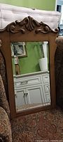 Antique wooden wall mirror with ornate carved top resting on a patterned fabric chair, reflecting room details.