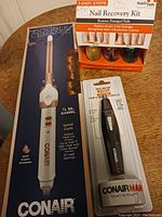 Photo showing Conair Double Ceramic curling iron, NailTek Nail Recovery Kit, and ConairMan nose/ear hair trimmer in packaging on a wooden surface.