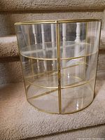 Curved glass display case with brass-colored metal frame and two shelves seen from the front on carpeted stairs
