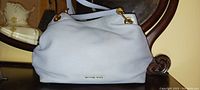 Front view of the Michael Kors handbag showing light blue leather, gold logo, and slouchy shape.