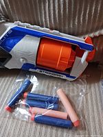 Close-up of the Nerf Strongarm blaster showing the white, blue, and orange body with included foam darts in a plastic bag under the blaster.