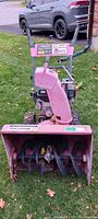 Front view of a used pink snowblower with control handles, chute, and flat tires, showing the working parts like the auger and engine.