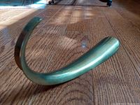 Angled side view of curved brass shoehorn on wooden floor showing smooth surface and patina.