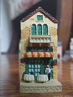 Close-up photo of a handcrafted cultured stone house featuring the 'BAILEY'S' sign, striped awning, and miniature outdoor setting with tables and figures