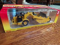 Side view of yellow and black Majorette construction scraper model in original box with clear plastic display window showing details of the vehicle.