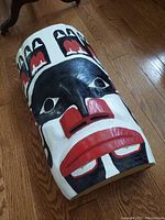 Hand carved wooden mask with stylized facial features, painted in black, white, and red, lying on a wooden floor