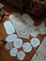 Photo showing 12 vintage handcrafted table doilies arranged on wooden floor with varying shapes and sizes including a long runner and several round and rectangular doilies
