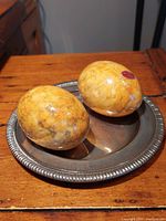 Two polished marble eggs placed on a silver-toned ornamental round dish atop a wooden surface, showing color and texture detail.