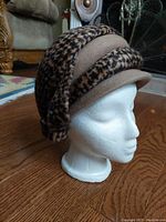 Side view of vintage mid 20th century ladies leopard print and beige layered fashion hat on white mannequin head with wooden floor background.