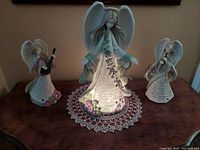 Set of three vintage illuminated festive musical angel figurines displayed on a wooden surface with a crochet doily beneath the largest angel, shown together for lot overview.