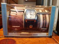 Front view of Fossil watch and three leather bands inside original clear plastic and cardboard box showing watch face and bands.
