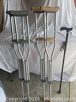 Two pairs of adjustable aluminum crutches, one with tan arm supports, the other with gray, and one black adjustable cane with wrist strap, all standing upright and leaning on a white surface.