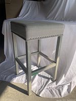 Grey wood bar stool with grey fabric upholstered square seat and nailhead trim, shown at a slight angle on a white backdrop.