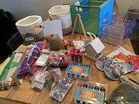 Full view of assorted kids art and craft supplies on a table including pom poms, paint pots, easels, wooden sticks, storage bins, and other craft materials.