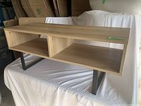 Angled view of the light brown wood veneer coffee table showing the top and front open cubby storage compartments with black metal legs.