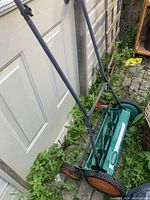 Photo showing manual push reel mower leaning against a white door outside, visible green frame, orange wheels, and black handles.