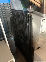 Folded or collapsed large black wire dog crate leaning against other items in storage.