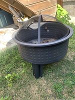 Fire pit shown with mesh spark screen cover placed on top, outdoors on grass near wooden shed.