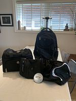 Photo showing all four bags on a table: large rolling backpack upright in back, two smaller bags and sports duffel in front