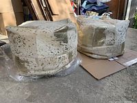 Two square fabric poufs with a cream base and gold accent patchwork design shown on garage floor with plastic wrapping and cardboard underneath.