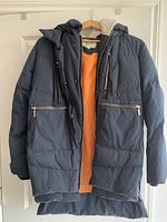 Navy blue Orolay winter jacket with orange inner lining and multiple zippered pockets hung on a wooden hanger against white door.