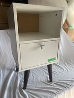 Front view of the white nightstand showing the cubby open shelf, drawer with metallic square handle, and dark legs.