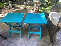 Two teal plastic folding side tables outdoors on stone patio. Both side tables are open and standing upright showing full shape and color.