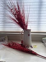 Photo of white metal jug holding red shiny decorative clusters and some jewelry on surface