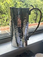 Side view of tall hammered silver metal pitcher with brass decorative leaf and twig handle, showing reflective hammered texture.