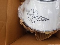 Top view of lamp shade showing black painted design on creamy white metal and plastic wrapping in shipping box